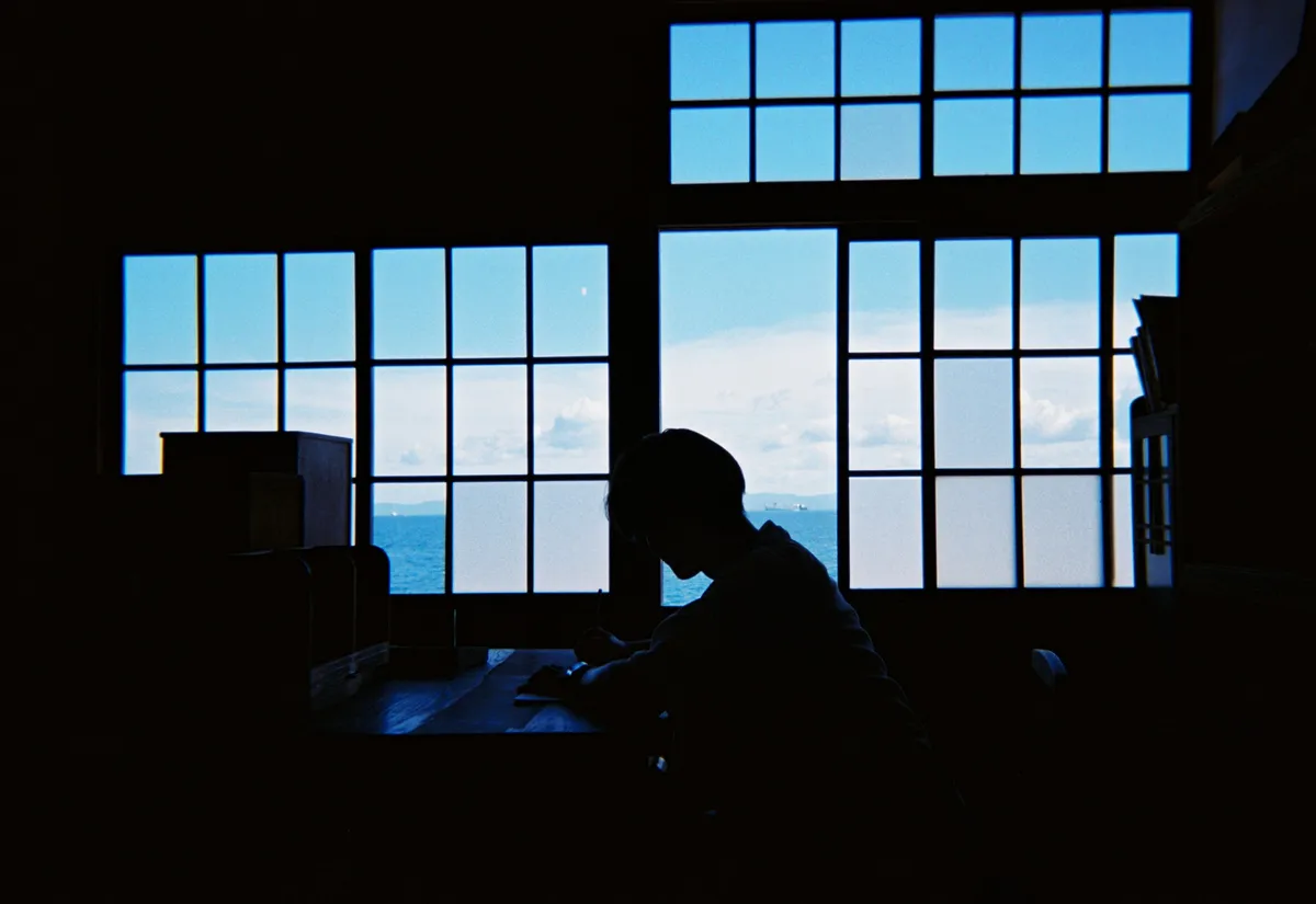 古い木枠の窓の前で、机に向かって書きものをする人。窓の外には海と空が広がっている